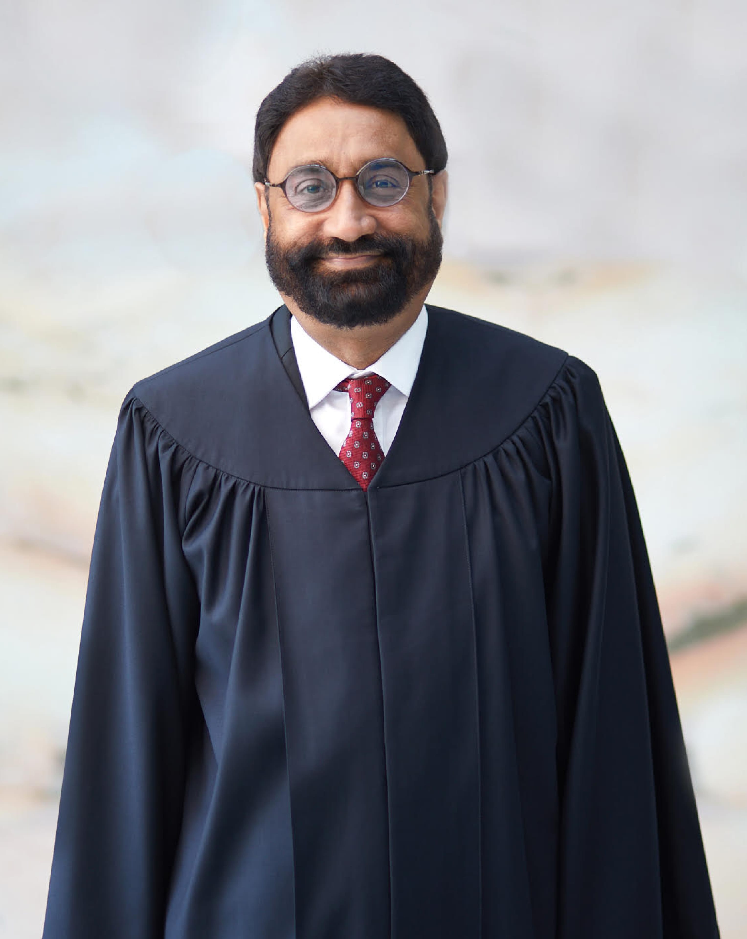Person with glasses, beard, in dark gown with red patterned tie.